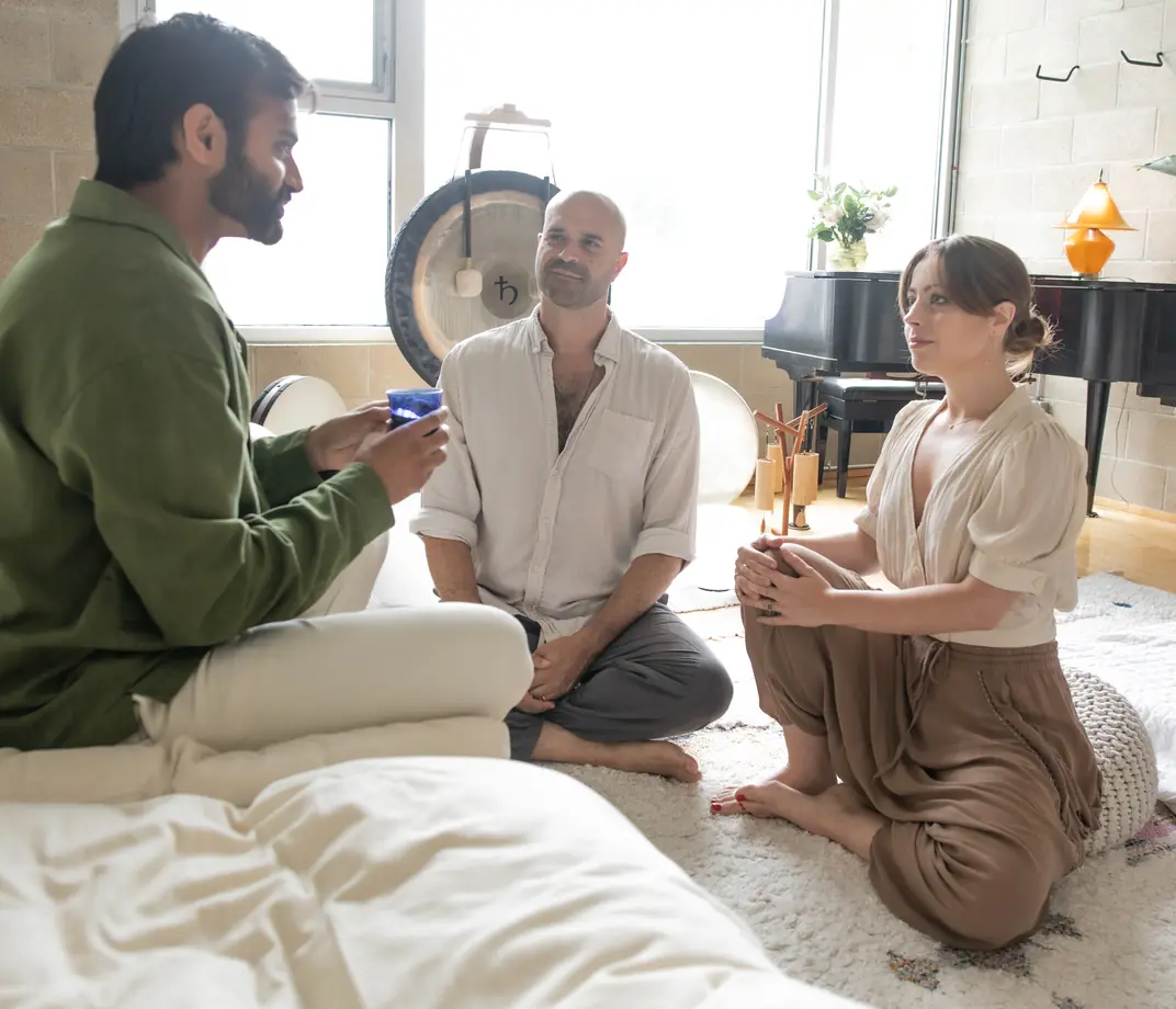 Yonatan sitting in conversation with two people over tea in the sunlit Word of Mouth studio, a gong and piano behind them