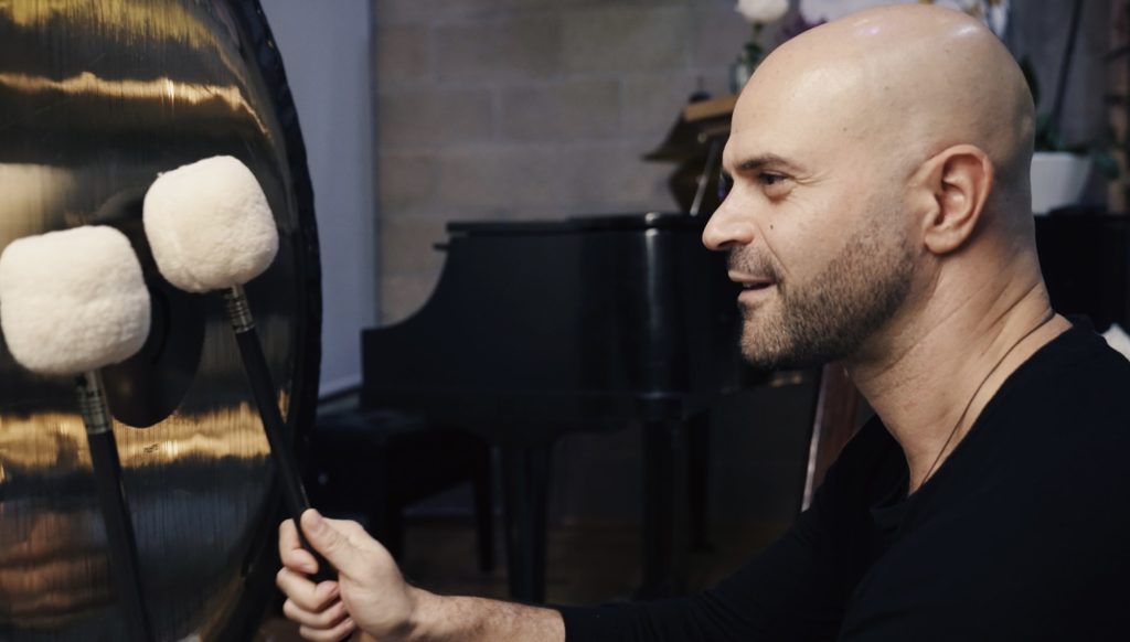 Yonatan Elkayam in profile beside the large hanging gong in the Sanctuary, holding two soft white mallets against it, the black baby grand piano behind him