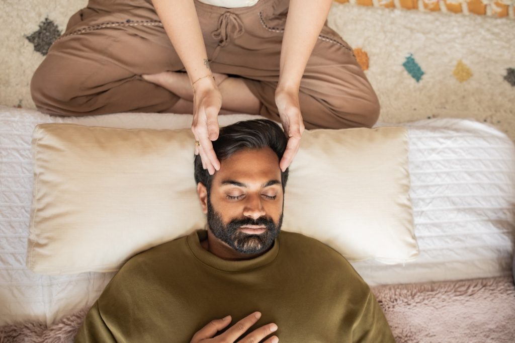 A man lying down with his eyes closed and one hand resting on his heart, receiving a Reiki session while the practitioner's hands hover just above his head.