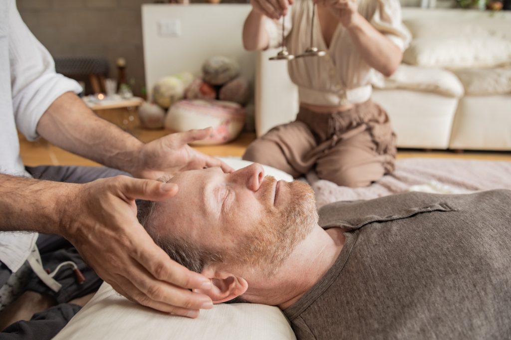 Yonatan Elkayam cradling a client's head with both hands during a session in the Word of Mouth loft, Jaime holding chimes above in the soft-focus background.