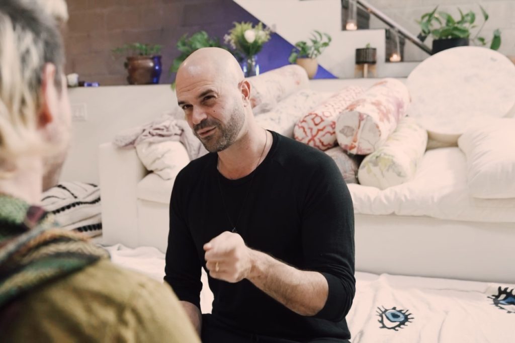 Yonatan Elkayam mid-sentence in a coaching session, seated on the floor of the Word of Mouth healing room across from a client.